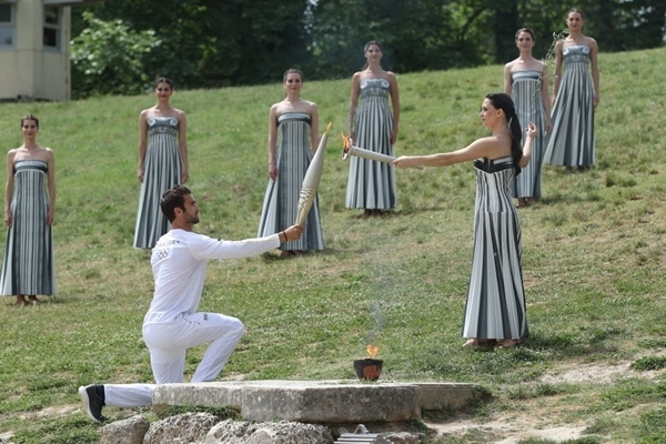 扮演最高女祭司的演員瑪麗·米娜（前右）點燃第一棒火炬手——希臘運動員、東京奧運會男子賽艇項目金牌得主斯特凡諾斯·恩圖斯科斯手中的火炬。新華社記者 趙丁喆 攝